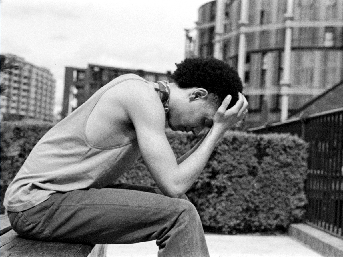 A young man sitting on a bench in a city park with his head in his hands
